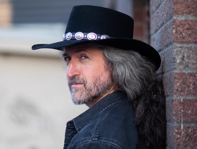 A photo of songwriter/musician/scholar Benton Roark, wearing a black hat and standing against a brick wall. (Photo credit: Joey Woolridge)
