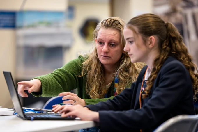 An STFC staff member helping a young female student with a task on a computer at a scientific computing schools day.