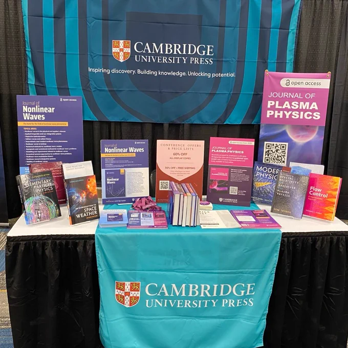 Display booth for Cambridge University Press at a conference, featuring banners and a variety of books related to physics, including titles on nonlinear waves, space weather, and plasma physics, as well as promotional material for the Journal of Plasma Physics and open access publishing.