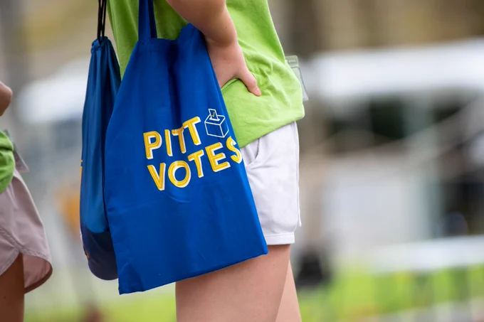 Close-up of a blue tote bag with the text 'PIT VOTES' in yellow, held by a person in a green shirt.