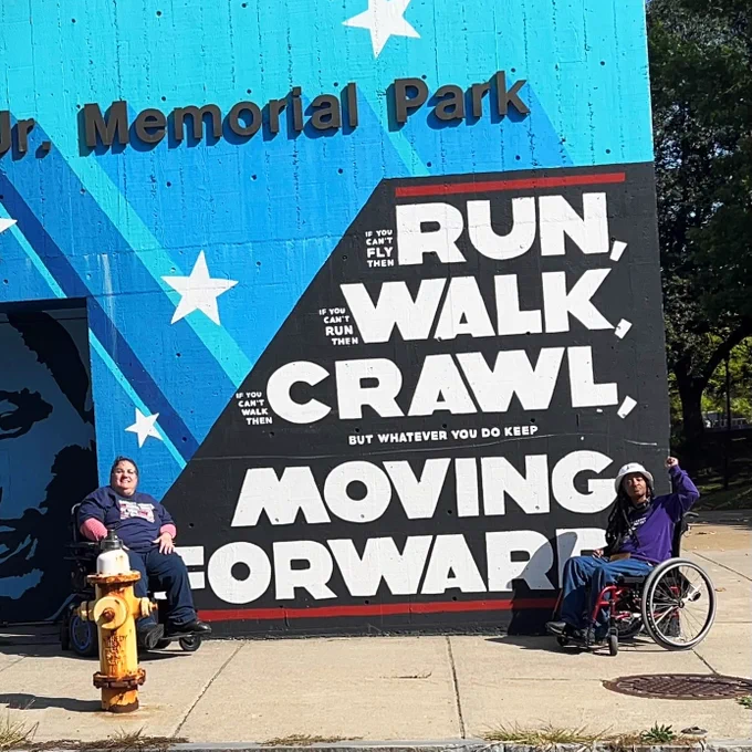 Susan Stahl and Anita Cameron sit in front of a painted sign at Martin Luther King Jr. Memorial Park. Susan is smiling, and Anita has one fist raised. The sign reads, "If you can't fly then run, if you can't run then walk, if you can't walk then crawl, but whatever you do keep moving forward."