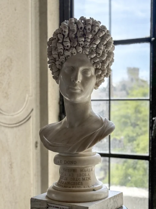 An incredibly elegant marble portrait bust of a Flavian woman. She has a square symmetrical face and high cheekbones atop an impossibly long swan-like neck. The bust reveals part of her shoulders, and her tunic and palla are worn low enough to expose a portion of her chest and right shoulder; the skin is very highly polished on her face. Her head is slightly canted and turned to her left. Her carved eyebrows meet in the middle of her nose, a mark of beauty at that time. What's most striking, however, is the towering hairstyle with its several rows of tight corkscrew curls. 