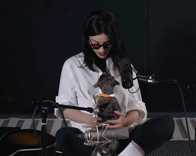 St. Vincent gently holds the grey puppy in her hands and close to her chest as the puppy looks up at her. she's looking down at the puppy as they share this moment.