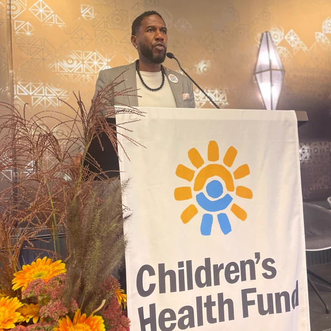 The Public Advocate speaks from a Children's Health Fund podium, flanked by flowers in autumnal colors