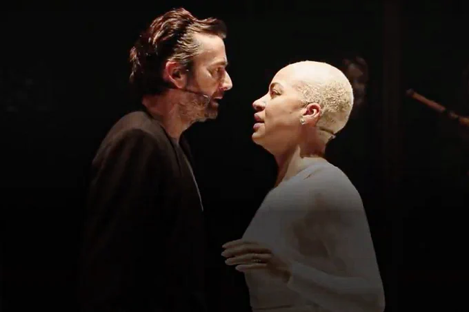 A photo shiowing two individuals in monochromatic clothes on a stage, looking worried at each other 