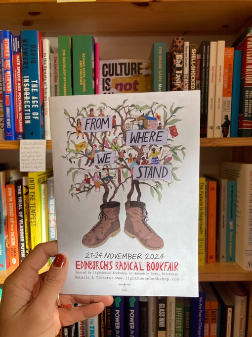 A brown hand with lovely red polished nails, holds up flyer of the Radical Book Fair program is held in front of a book case of leftie politics titles - it is illustrated with two boots from which trees grow and in the branches sit activists and readers and a richly diverse community holding banners that read 'No justice no peace and "Another World Is Possimble' and in big letters the theme for the fair 'From Where We Stand'.