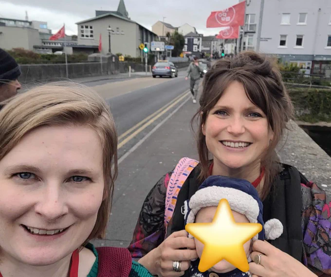Our Tours Producer Alison and Artistic Director Ellie smile into the camera for a selfie. They are standing on a bridge, with the river visible to their right, and buildings behind them. Ellie’s baby is in a baby carrier on her front. The baby’s face is covered with a star emoji. In the background, the streets are lined with red flags for Babaro Festival.