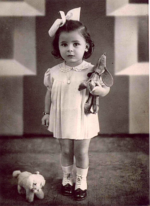 Vintage black and white photo of a young girl holding a toy and standing next to a stuffed animal, set against a patterned backdrop.