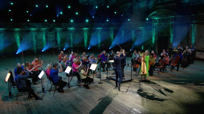 An orchestra in casual colourful outfits playing in a dramatically lit hall