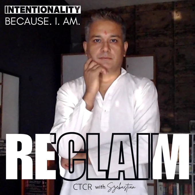 Szebastian standing in their work-studio, sporting white outfit, salt and pepper short spiked hair and looking straight into the camera with a thoughtful gaze. 