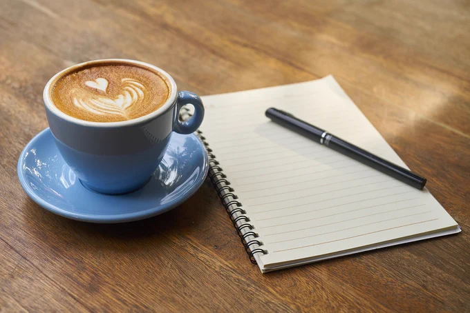 A notebook, a black pen and a blue cup and saucer with a latte in.