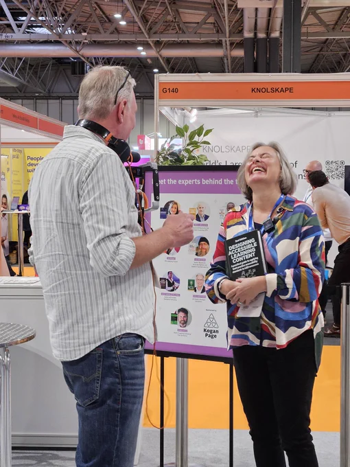 A man and a lady having a conversation. The lady is laughing. The man has a set of headphones around his neck. Behind them is a billboard advertising the Meet the author session at World of Learning 