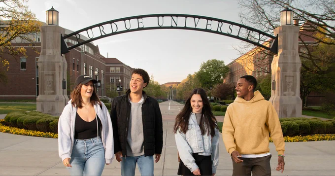 Four students smiling and walking under the Purdue University gateway arch during a sunny day.