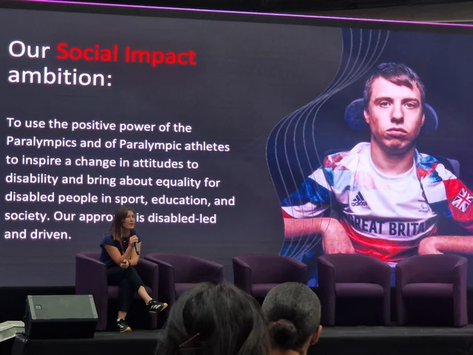 Anna sitting on stage with large image of Paralympian in background and words "our social impact ambition" - talking about changing attitudes through sport.