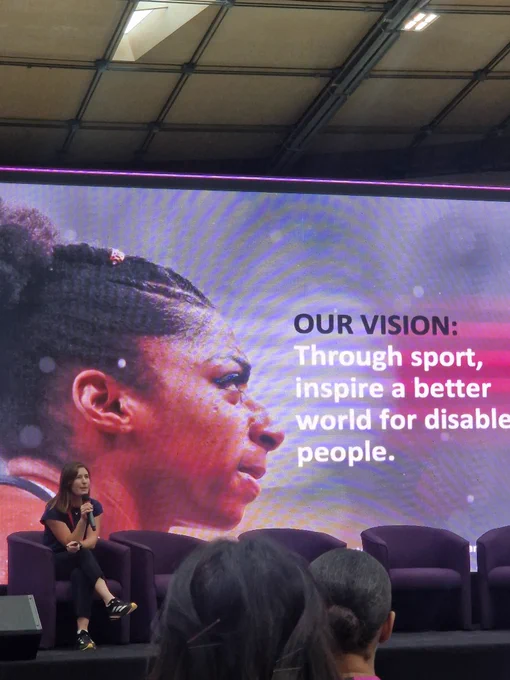 Anna on stage at event, with large image of athlete and words "our vision. Through sport, inspire a better world for disabled people"