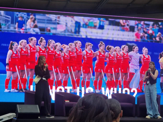 Tess & Suzy, on stage, with image of Suzy competing at Paris 2024 alongside GB hockey team