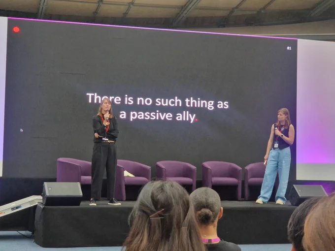 Tess and Suzy, in discussion on stage, with background "there is no such thing as a passive ally"