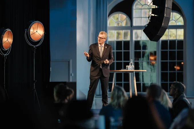 Full-color photo of David speaking to an audience onstage, with lights off to the side and people gathered in the foreground.