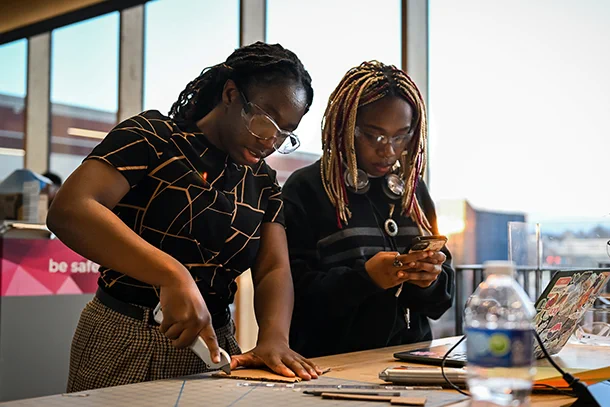Two students collaborate on an engineering project, one is cutting cardboard with a box cutter and one is looking at a cell phone