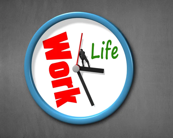 Clock with "Work" written in red taking up more than half of the face of the clock and "life" in green taking up about a fourth, while a man stands on the hands at 3:30 trying to push back the work section