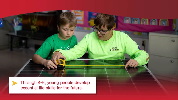 Two young individuals in 4-H t-shirts are engaged in measuring a project on a table, illustrating a hands-on educational activity.