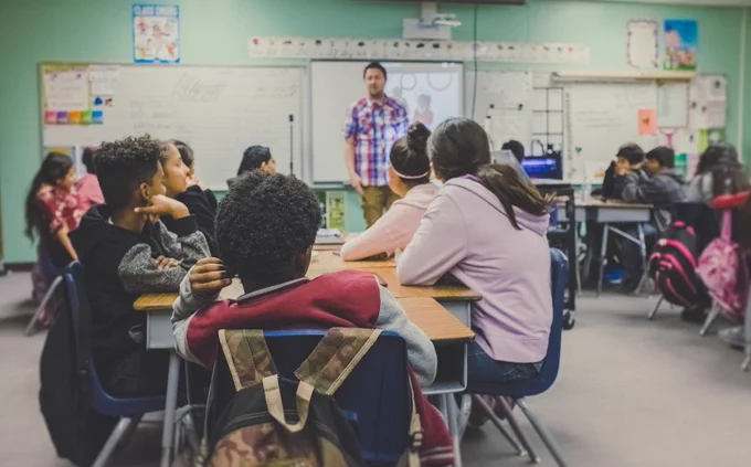 Students sitting at desks in a classroom. A teacher is standing in front of them.