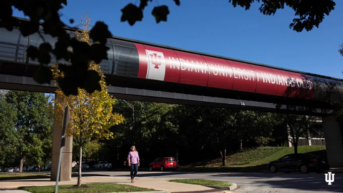 A person walks on a sidewalk near a road, beneath an overpass featuring a large banner that reads "INDIANA UNIVERSITY INDIANAPOLIS" with the university's logo. Sunlight filters through trees surrounding the area.