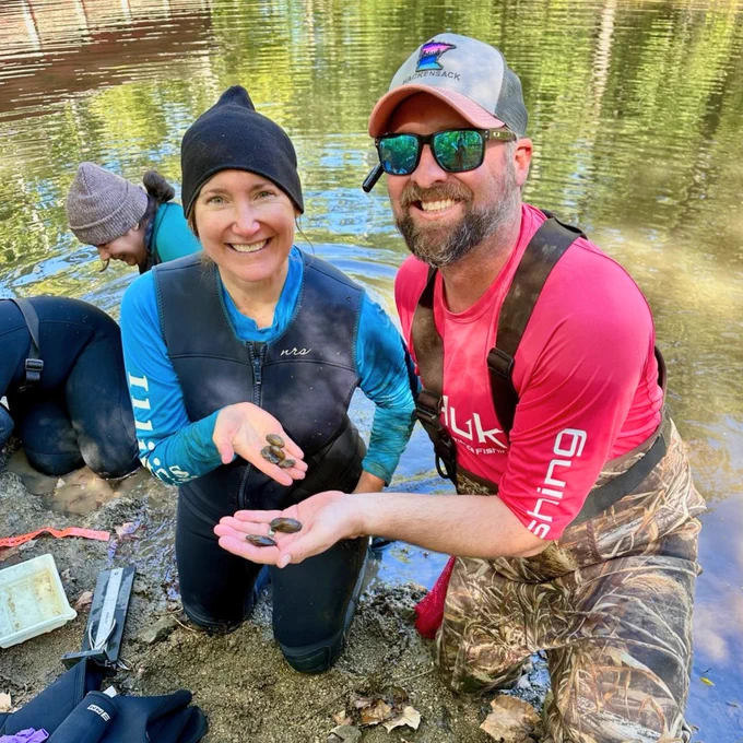 A woman and man standing along a river bank holding salamander mussels and mussel shells.