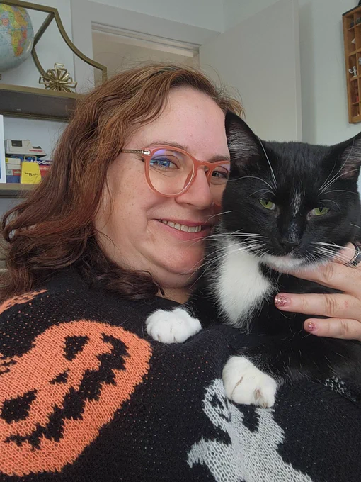 A woman in a black cardigan with an orange pumpkin and white ghost on it holding a tuxedo cat