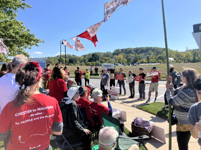 A crowd of people standing and sitting outside, some holding signs or banner poles, across the street from a sign that says “Vanguard”