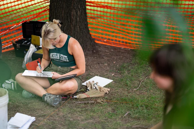 Woman writing on a clipboard in a wooded area with tools and an orange safety fence around her.
