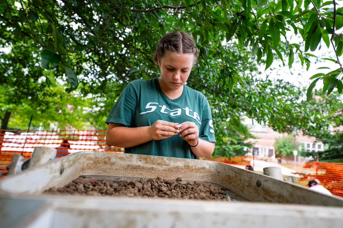 A young woman examines an object over a dirt-filled sifting screen in a wooded outdoor area.
