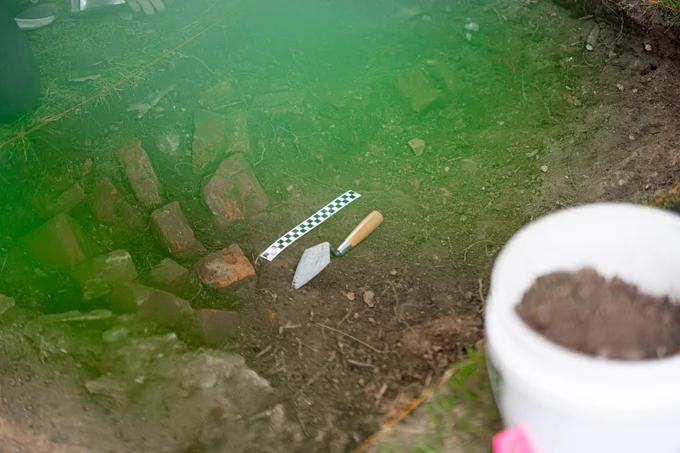 An archaeological dig site with exposed bricks, a trowel, and measuring tape.