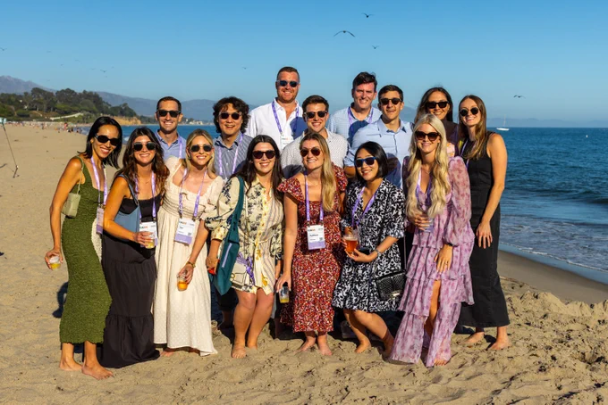 Group of professionals on a beach, wearing conference badges, posing for a photo, with the ocean in the background.