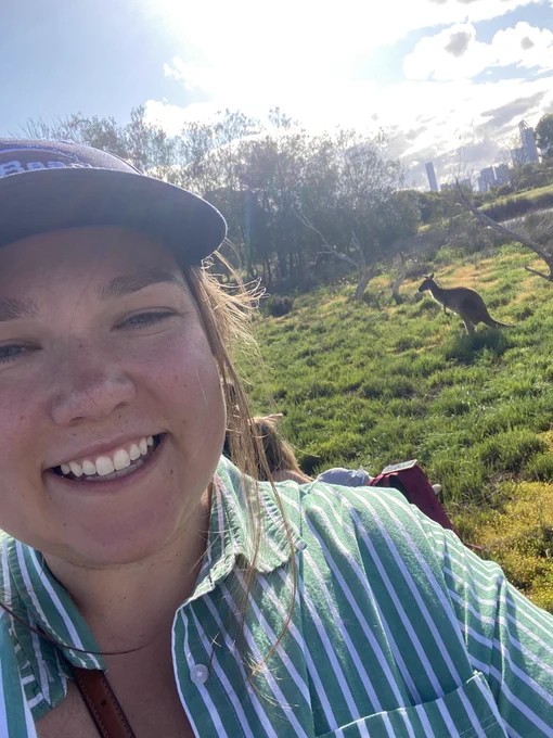 Selfie of Sydney wearing a baseball hat and striped shirt. There is a Kangaroo standing in the background.