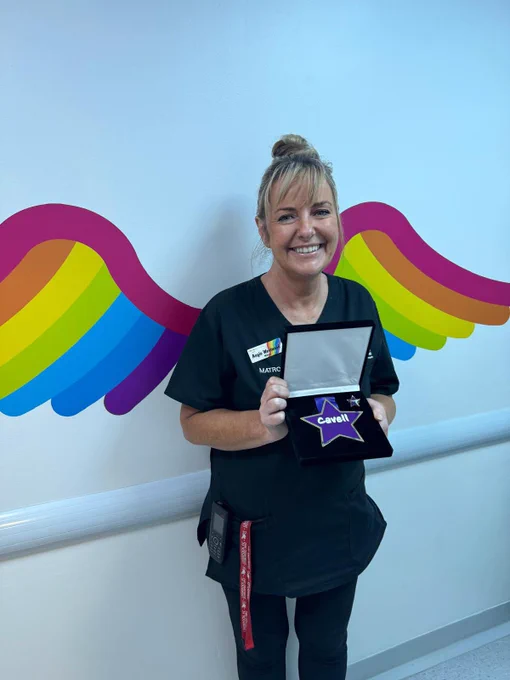 Lady in matron uniform holding an award in front of some multicoloured Angel Wings