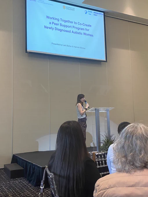 Woman holding microphone presenting in front of slide that reads “working together to co-create a peer support program for newly diagnosed autistic women”