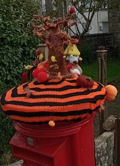 A yarn bombed post box. The top is covered in a giant knitted orange and black striped "beret" with a small macrame tree growing out of the centre. A gnome is sitting on a swing hanging from one of the branches, and there are various fabric pumpkins around the base