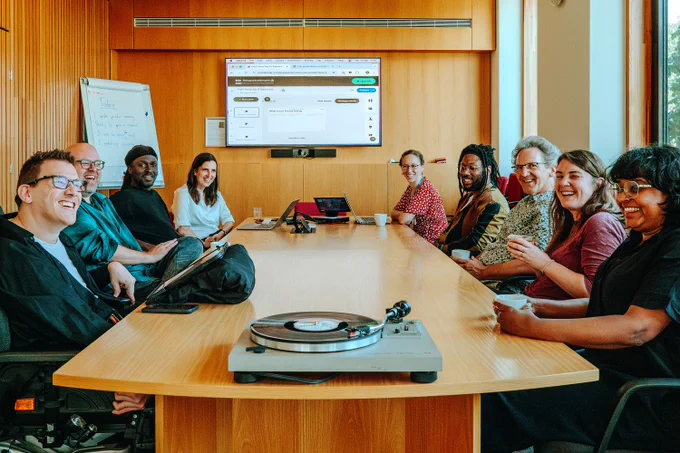 The Hub’s Hub Prioritisation and Selection Committee sitting around a conference table in a meeting room. They are smiling and appear to be engaged in a relaxed, collaborative discussion. On the table in the foreground, there is a record player. The room is warm and well-lit, featuring wood paneling and a whiteboard, and a large screen is visible at the far end of the room.