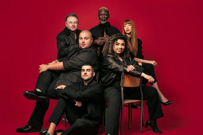 A photo showing a group of six performers arranged on chairs, dressed in dark clothing and striking a sultry pose in front of a red backdrop 