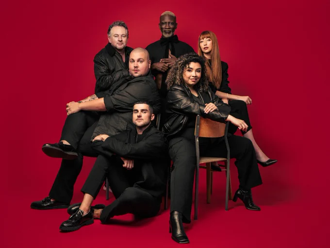 The cast of Kinky Boots (2025), stood and sat around chairs - all dressed in black on a red background, looking into camera.