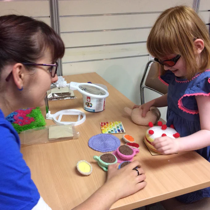 a woman in a bright blue t-shirt sitting at a table playing kitchens with a little girl who is sitting opposite wearing a blue dress and dark glasses