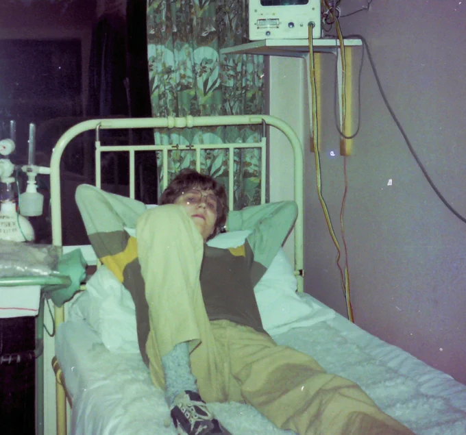 Photo of Bert as a teenager lying on a bed at Harefield Hospital. He is wearing cream trousers and a brown and cream top.