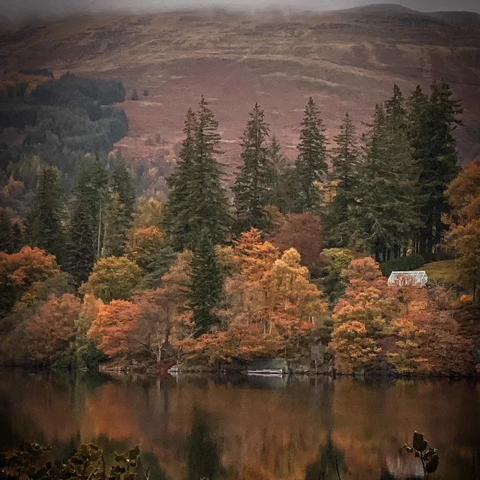 Autumn foliage surrounding a small cottage and a boat on the shore of a serene loch, with misty hills in the background. Picture taken by @steviesorefingers on Instagram