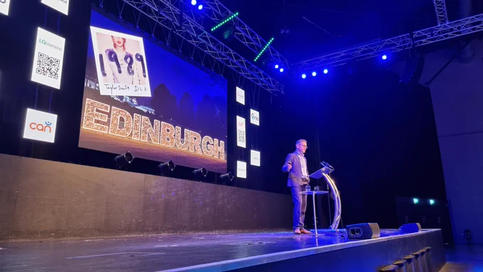 Mike Pinkerton stands on stage looking out at the Comms Academy audience, behind him a large screen featuring a Taylor Swift album cover and the word EDINBURGH in gold lights.