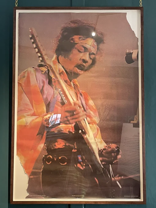 Framed photograph of Jimi Hendrix, a Black man with mid-length black hair, orange scarf bandana and matching kimono jacket, playing electric guitar. The image is set against a teal green wall.