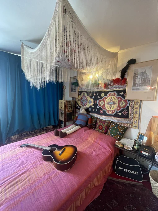 Photograph of Jimi Hendrix’s London apartment bedroom with red and orange bedspread, tapestries hung on wall for decoration and blue curtains across window. On the bed is a large knitted dog character in red and blue jumper, alongside an acoustic guitar.