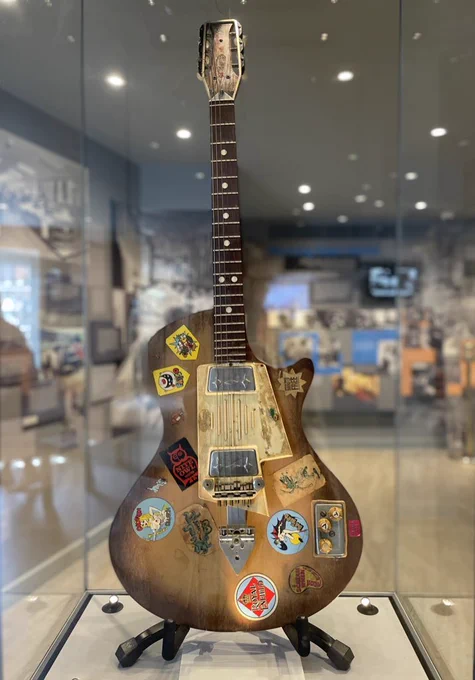 Photograph of Jimi Hendrix’s dark brown wood effect guitar in glass case. The guitar is covered with various stickers.