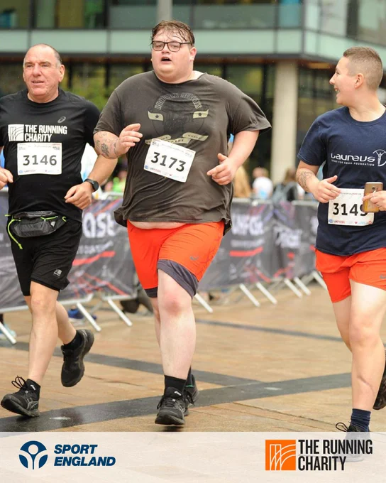 Three participants in a public running event, wearing numbered bibs and sportswear, jogging on a city street with event banners visible in the background.