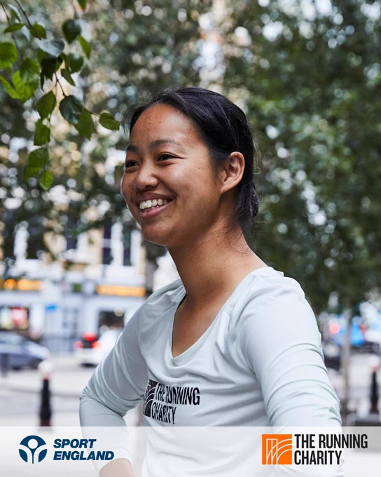 Person smiling in a white t-shirt with logo of The Running Charity, standing outdoors among trees.